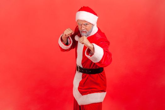 Strict elderly man with gray beard wearing santa claus costume standing with fists and being ready to fight, looking at camera with angry expression. Indoor studio shot isolated on red background.