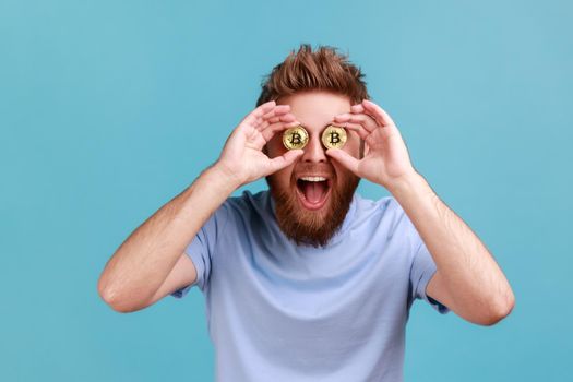 Bitcoin, digital money. Portrait of playful excited bearded man covering eyes with golden bitcoins, being confident in cryptocurrency. Indoor studio shot isolated on blue background.
