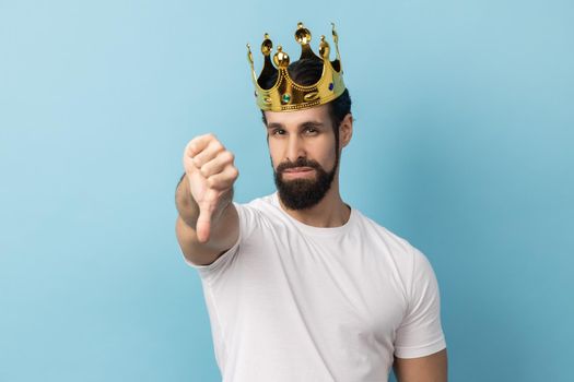 Portrait of sad serious man with beard in white T-shirt and in gold crown standing looking at camera with disappointed expression, showing thumb down. Indoor studio shot isolated on blue background.