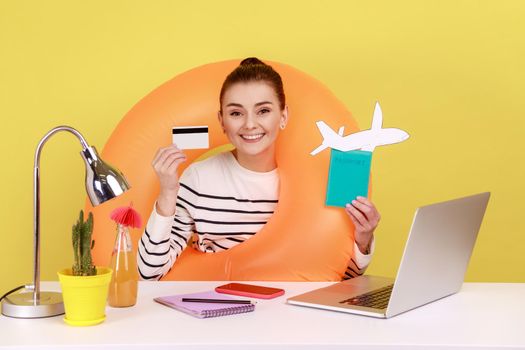 Smiling happy woman with rubber ring holding and showing credit card and passport, booking tickets and hotel online on laptop. Indoor studio studio shot isolated on yellow background.
