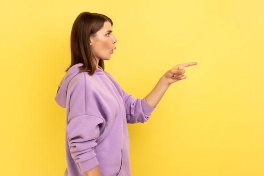 Side view of excited beautiful young adult woman standing and pointing at copyspace with surprised face, wearing purple hoodie. Indoor studio shot isolated on yellow background.