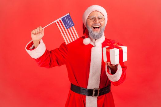 Portrait of excited elderly man with gray beard wearing santa claus costume holding in hands USA flag, giving present box for winter holidays. Indoor studio shot isolated on red background.