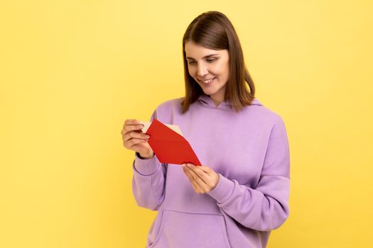 Side view of smiling young woman opening envelope and reading letter, with positive expression, got unexpected pleasant news, wearing purple hoodie. Indoor studio shot isolated on yellow background.