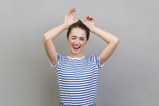 Portrait of playful woman wearing striped T-shirt making funny bunny ears with hands on head, childish behavior, optimistic mood. Indoor studio shot isolated on gray background.