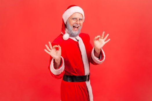 Portrait of happy elderly man with gray beard wearing santa claus costume with smile making ok gesture being agreeable, showing okay sign. Indoor studio shot isolated on red background.