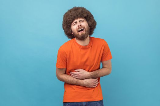 Stomach pain. Portrait of sick sad unhealthy man with Afro hairstyle wearing orange T-shirt standing and holding his painful belly. Indoor studio shot isolated on blue background.