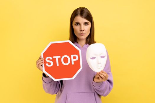 Portrait of serious woman holding red stop sign and white mask, looking at camera with strict bossy expression, wearing purple hoodie. Indoor studio shot isolated on yellow background.