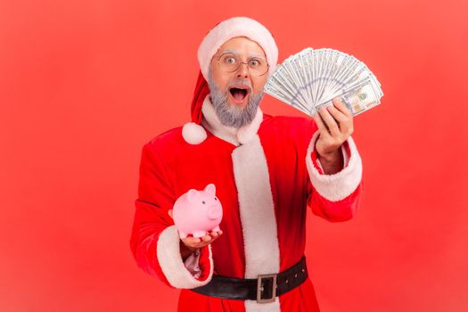 Surprised elderly man with gray beard wearing santa claus costume holding dollars banknotes and piggy bank, savings, high rate deposit. Indoor studio shot isolated on red background.