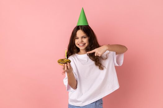 Little girl in funny party cone on head pointing at cake with candle and looking at camera with toothy smile, expressing pure child happiness. Indoor studio shot isolated on pink background.