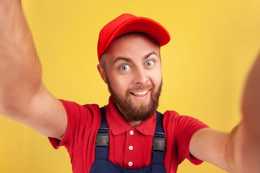 Optimistic happy man with wearing red cap and blue uniform taking selfie, looking at camera funny facial expression POV, point of view of photo. Indoor studio shot isolated on yellow background.