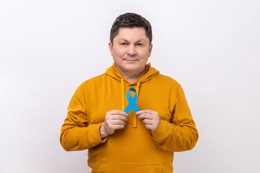 Prostate cancer awareness. Serious man holding blue ribbon in his hands, supporting people living and illness, wearing urban style hoodie. Indoor studio shot isolated on white background.