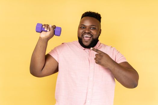 Portrait of excited positive bearded man wearing pink shirt standing with dumbbell and pointing at sport equipment, looking at camera. Indoor studio shot isolated on yellow background.