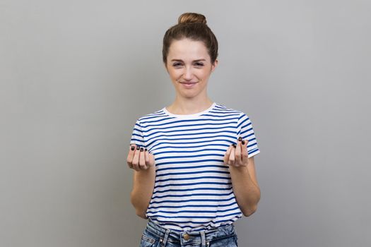Portrait of attractive positive woman wearing striped T-shirt smiling and showing give me money gesture, asking for payment, allowance. Indoor studio shot isolated on gray background.
