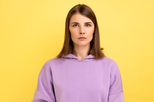 Portrait of young serious unsmiling strict woman looking at camera with calm attentive focused expression, wearing purple hoodie. Indoor studio shot isolated on yellow background.