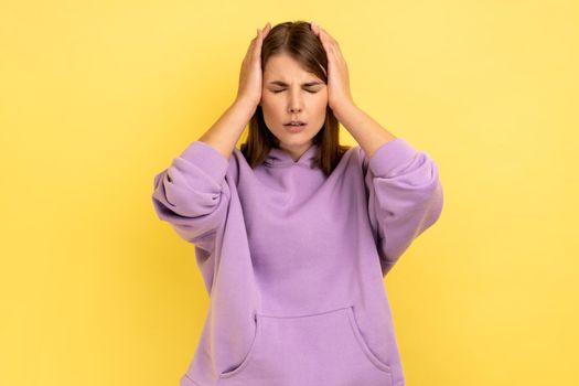 Portrait of ill woman clasping hands over sore head, frowning from acute pain suffering unbearable migraine, stress tension, wearing purple hoodie. Indoor studio shot isolated on yellow background.