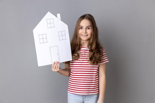 Portrait of charming little girl wearing striped T-shirt holding paper house and smiling friendly, advertising realtor services, house purchase. Indoor studio shot isolated on gray background.