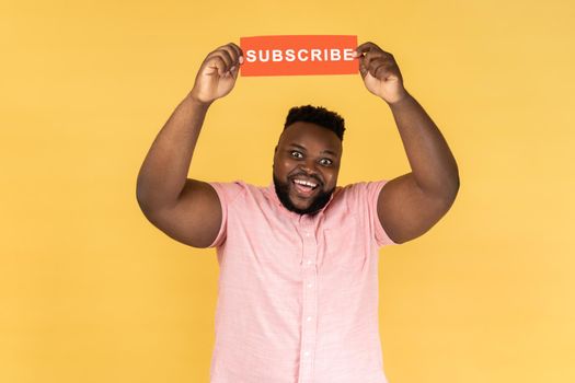 Portrait of excited amazed man in pink shirt holding with paper card with subscribe inscription above head, looking at camera with toothy smile. Indoor studio shot isolated on yellow background.