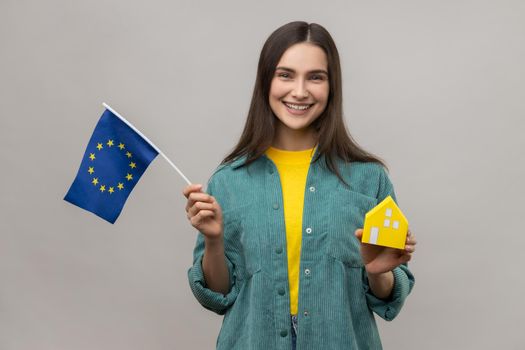 Portrait of positive satisfied holding european flag and paper house, dreaming to buy accommodation in Europe, wearing casual style jacket. Indoor studio shot isolated on gray background.
