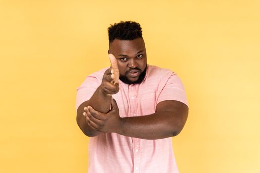 I'll kill you. Portrait of man wearing pink shirt pointing finger guns to camera and looking aggressive, threatening to shoot, hands imitating weapon. Indoor studio shot isolated on yellow background.