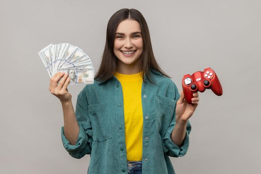 Portrait of extremely happy woman showing to camera red joypad and dollars banknotes, playing and earning money, wearing casual style jacket. Indoor studio shot isolated on gray background.