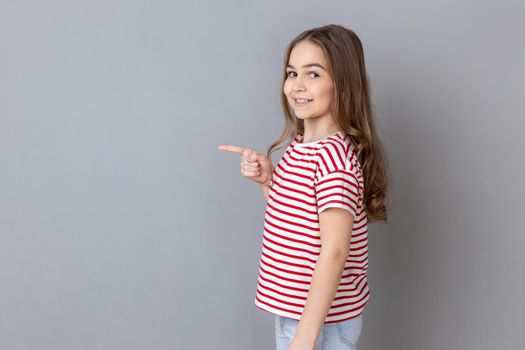 Wow, look at crazy advertisement. Side view of little girl wearing striped T-shirt pointing aside at empty place for ad content and expressing happiness. Indoor studio shot isolated on gray background