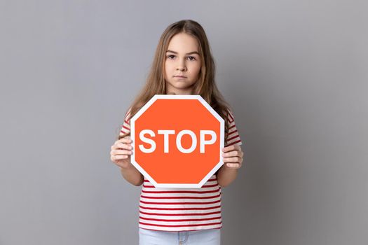 Portrait of serious strict little girl wearing striped T-shirt holding red stop sign, looking at camera with serious facial expression, prohibition. Indoor studio shot isolated on gray background.
