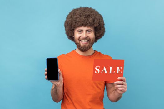 Portrait of satisfied man with Afro hairstyle wearing orange T-shirt holding smartphone with blank display and sale card in hands, discount application. Indoor studio shot isolated on blue background.