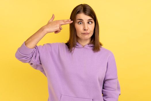 Portrait of desperate woman, pointing finger gun to head, shooting herself, making suicide gesture to stop depression, wearing purple hoodie. Indoor studio shot isolated on yellow background.