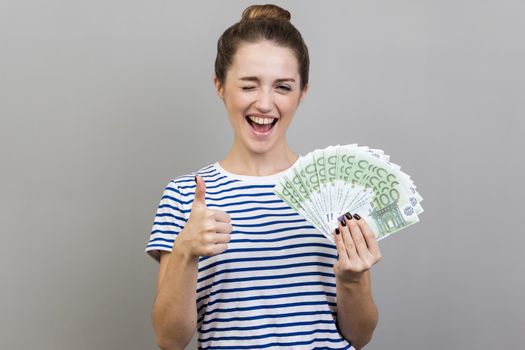 Portrait of woman wearing striped T-shirt holding bunch of euro banknotes and winking, showing thumbs up gesture, sign of success, approval. Indoor studio shot isolated on gray background.