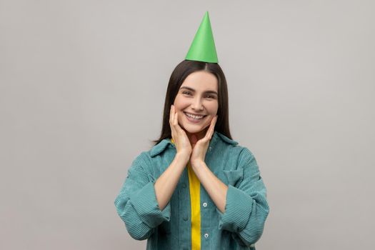 Beautiful woman with dark hair in birthday party cone and smiling to camera, enjoying birthday celebration, festive mood, wearing casual style jacket. Indoor studio shot isolated on gray background.