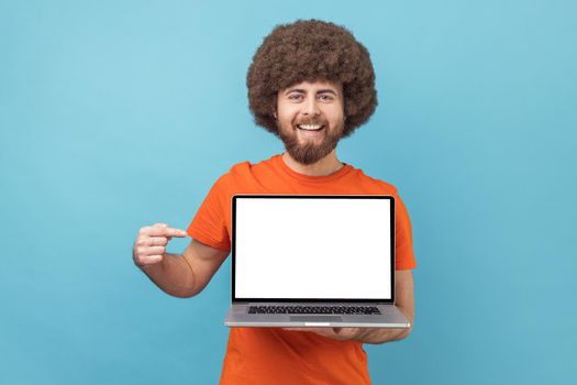 Portrait of delighted man with Afro hairstyle wearing orange T-shirt standing showing empty display of laptop, pointing at screen, advertising area. Indoor studio shot isolated on blue background.
