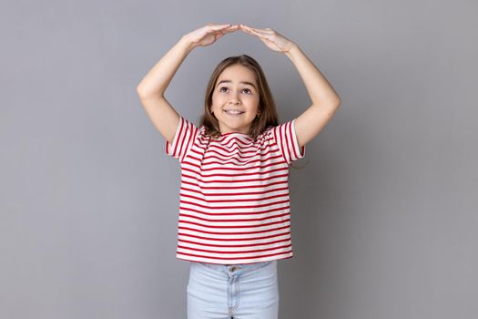 I am in safety. Portrait of little girl wearing striped T-shirt making house roof gesture and smiling, concept of life insurance, protection. Indoor studio shot isolated on gray background.