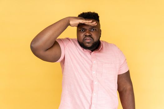 Portrait of curious man with beard in pink shirt keeping palm over head and looking attentively far away, peering with expectation at long distance. Indoor studio shot isolated on yellow background.