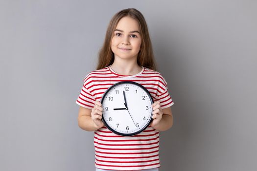 Portrait of smiling little girl wearing striped T-shirt holding big wall clock, looking at camera with pleasant emotions, time to go. Indoor studio shot isolated on gray background.