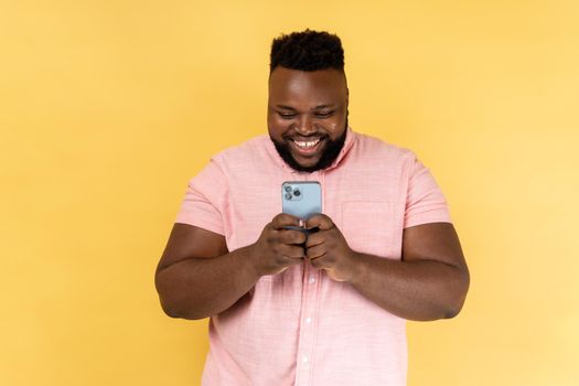 Portrait of bearded happy man wearing pink shirt using mobile phone with happy expression, addicted to smartphone, texting in social network. Indoor studio shot isolated on yellow background.