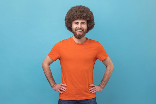 Portrait of cheerful man wearing orange T-shirt looking at camera with toothy smile and happy facial expression, being in good mood, keeps hands on hips. Indoor studio shot isolated on blue background