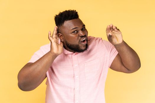 Portrait of unsure bearded young adult man wearing pink shirt standing with hand near ear, listening with frowning face, showing blah blah gesture. Indoor studio shot isolated on yellow background.