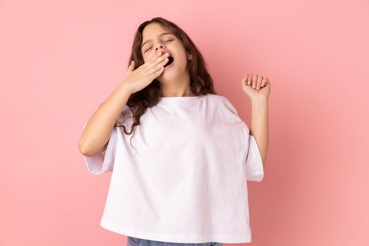 Portrait of adorable sleepy little girl wearing white T-shirt standing and yawning with closed eyes and raised arm, covering mouth with palm. Indoor studio shot isolated on pink background.