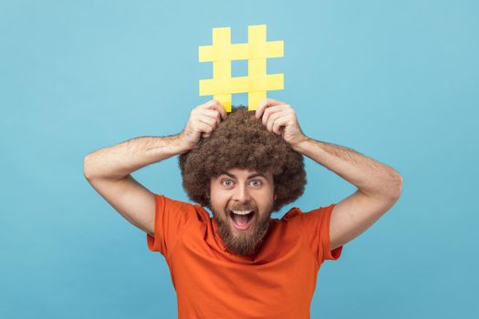 Portrait of man with Afro hairstyle holding yellow hashtag sign board on head, dreaming about target tagging posts on his website, smm manager. Indoor studio shot isolated on blue background.