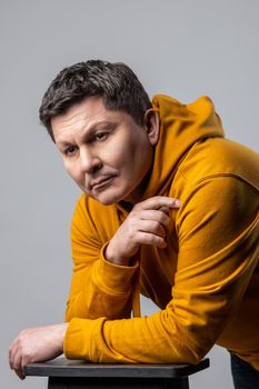 Portrait of handsome middle aged man looking pensive thoughtful, leaning on chair, being deep in thoughts, wearing urban style hoodie. Indoor studio shot isolated on gray background.