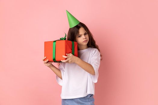 Portrait of curious little girl wearing white T-shirt and party cone holding red wrapped present box, being interested white inside of gift. Indoor studio shot isolated on pink background.