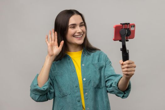 Attractive woman holding smartphone on steadycam in hand, blogger broadcasting livestream, waving hand, wearing casual style jacket. Indoor studio shot isolated on gray background.