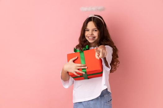 Little girl wearing white T-shirt with holy nimbus with kind smile holding gift box, charity organization, pointing to camera with finger, religion. Indoor studio shot isolated on pink background.