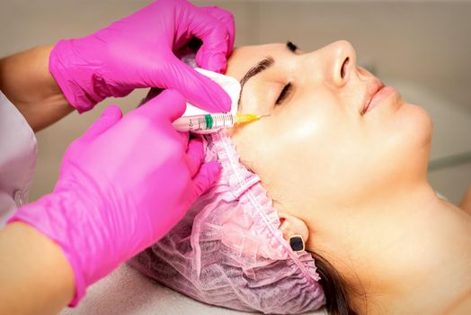 The young white woman is getting rejuvenating facial injections with hyaluronic acid on the eye in a beauty clinic