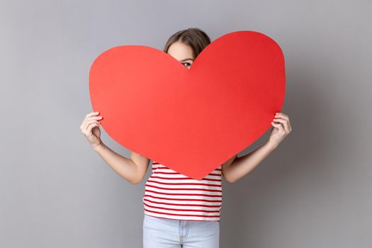 Portrait of adorable little girl wearing striped T-shirt peeking from big red heart and expressing positive emotions, falling in love. Indoor studio shot isolated on gray background.