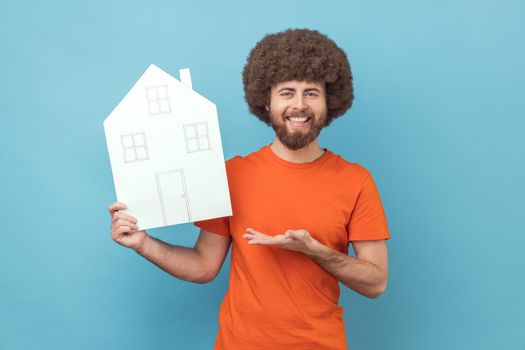 Man with Afro hairstyle presenting paper house with drawn windows and doors, making project of home of her dreams, looking at camera with smile. Indoor studio shot isolated on blue background.