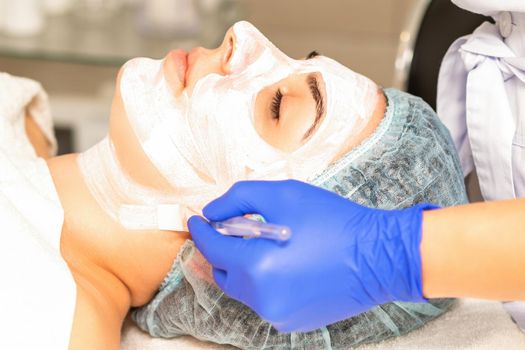 The beautician with brush applies a photochemical and glycolic peeling face mask to the female patient face in the beauty clinic