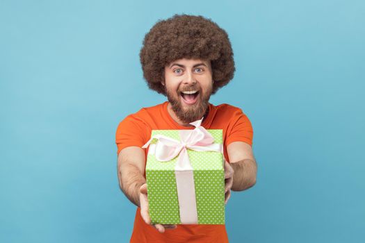 Portrait of man with Afro hairstyle wearing orange T-shirt giving gift, congratulating on birthday and offering surprise, holiday bonus wrapped in box. Indoor studio shot isolated on blue background.
