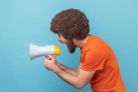 Side view of man with Afro hairstyle wearing orange T-shirt loudly screaming at megaphone, making announce, protesting, wants to be heard. Indoor studio shot isolated on blue background.