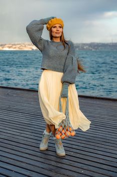 Outdoors fashion portrait of a beautiful middle aged woman walking on the beach. Marine background. Dressed in a stylish warm blue sweater, yellow skirt and beret